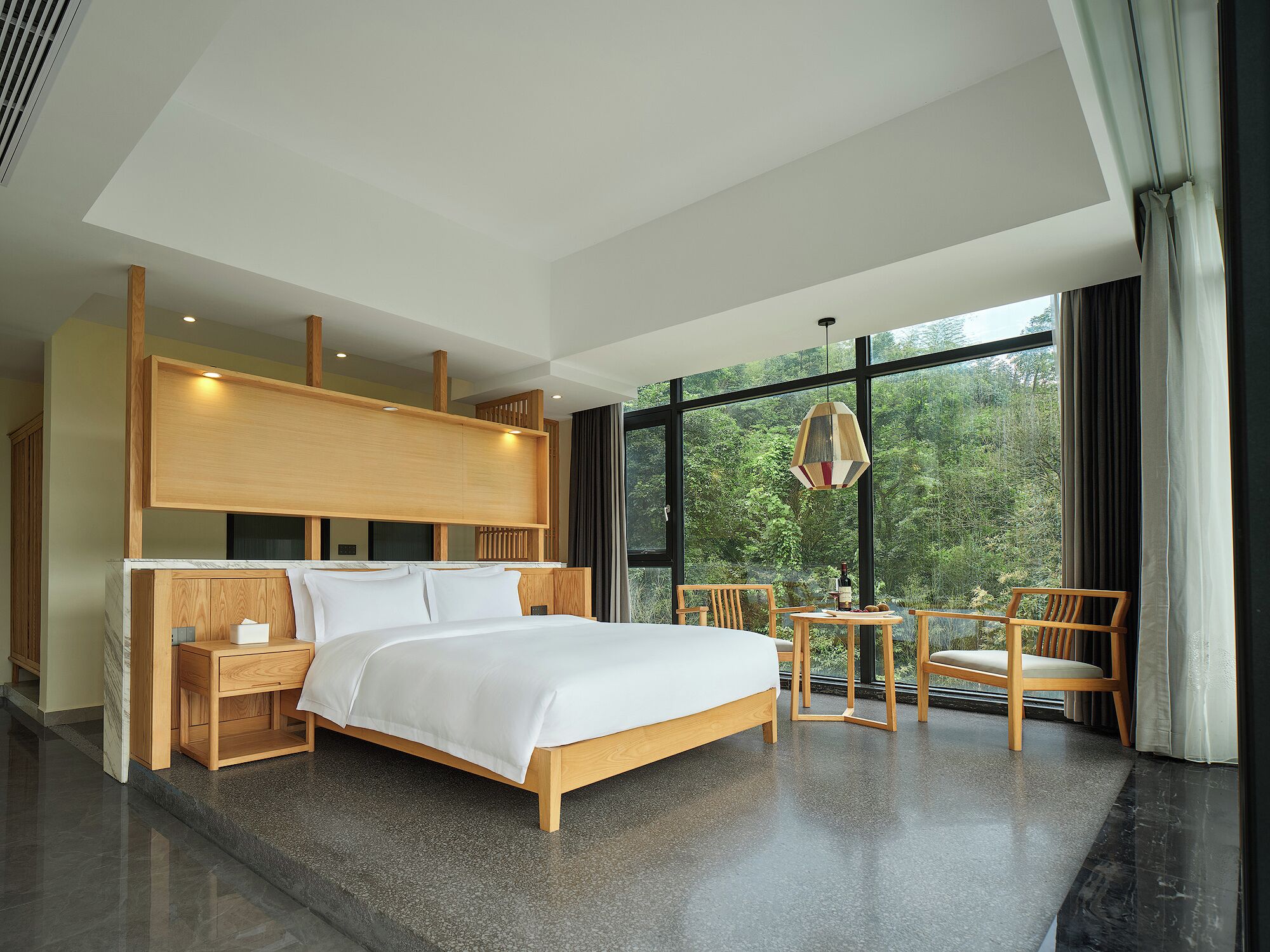 Une chambre d’hôtel moderne avec un grand lit, une tête de lit en bois, une petite table avec des chaises et une vue sur la forêt grâce à de grandes fenêtres.