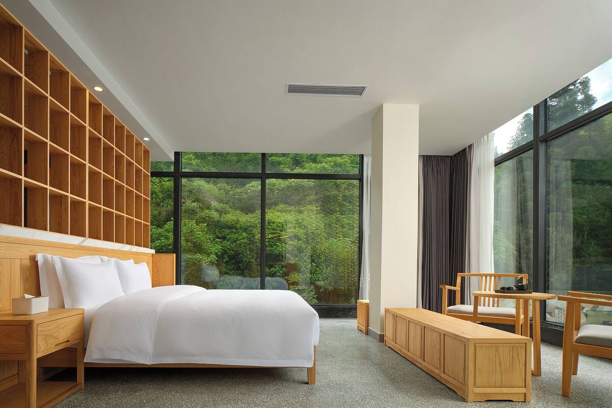 A modern hotel room with a large bed, wooden headboard, and a view of lush greenery through large windows.