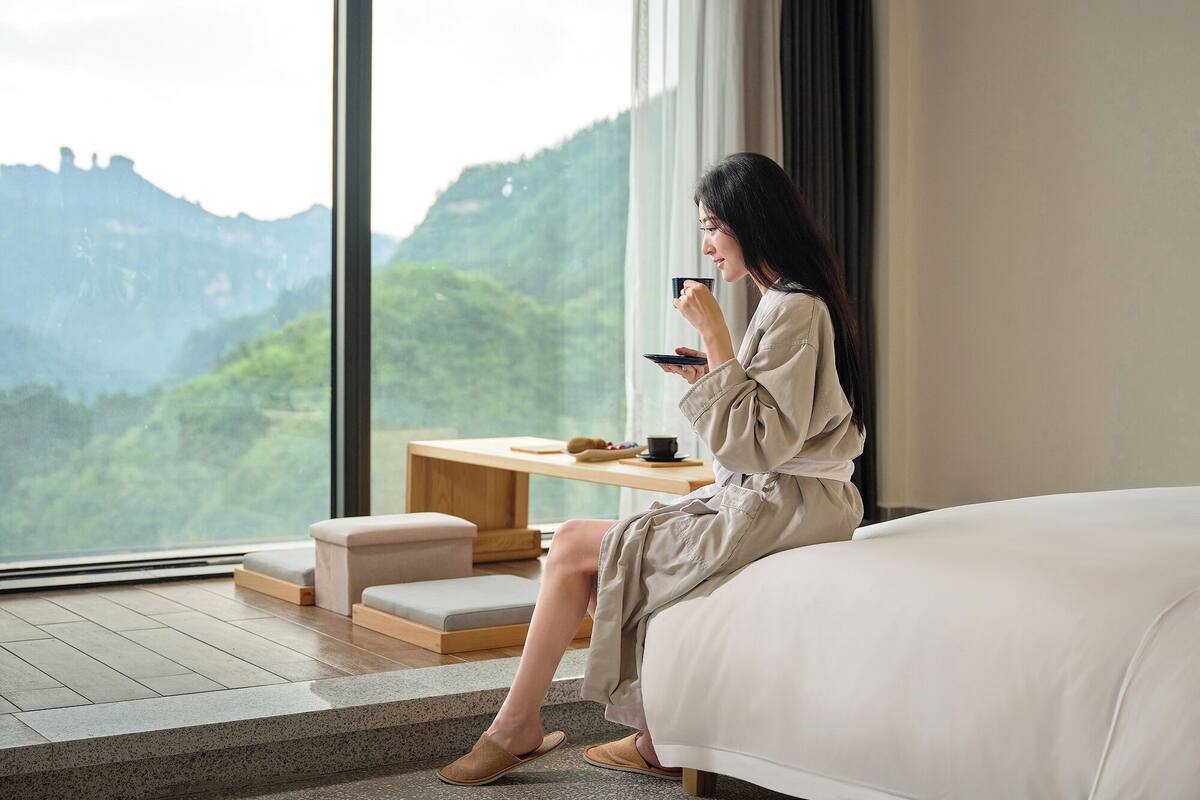 A woman sitting by a window in a room with a bed, a small table, and a view of a mountainous landscape.
