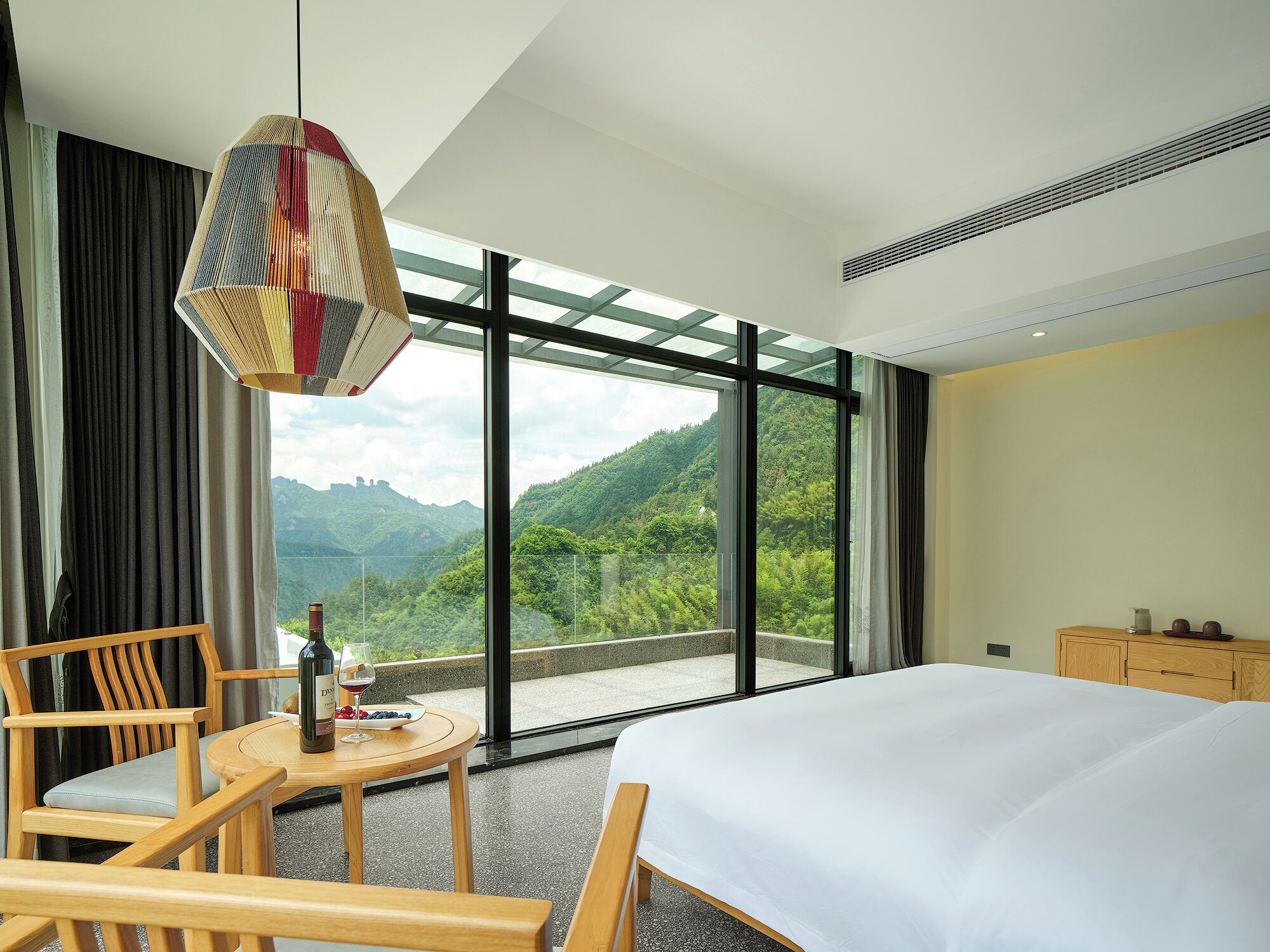 Hotelkamer met een groot bed, een houten stoel, een klein rond tafeltje met fles en glas, en uitzicht op een bergachtig landschap door grote ramen.