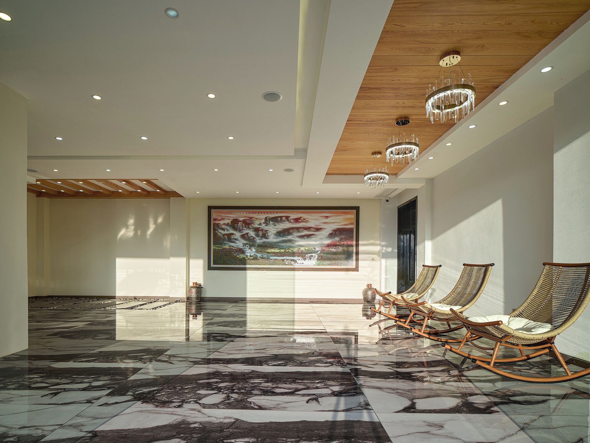 A spacious lobby with a marble floor, wooden ceiling, and a large painting. There are wicker rocking chairs and a large vase on the floor.