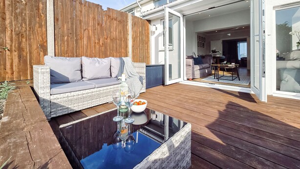Une terrasse en bois avec un canapé en osier, une table en verre surmontée de verres à vin et un saladier rempli de fruits.
