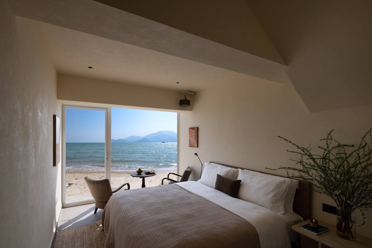 A bedroom with a large bed, a chair, a small table, and a view of the beach and ocean.