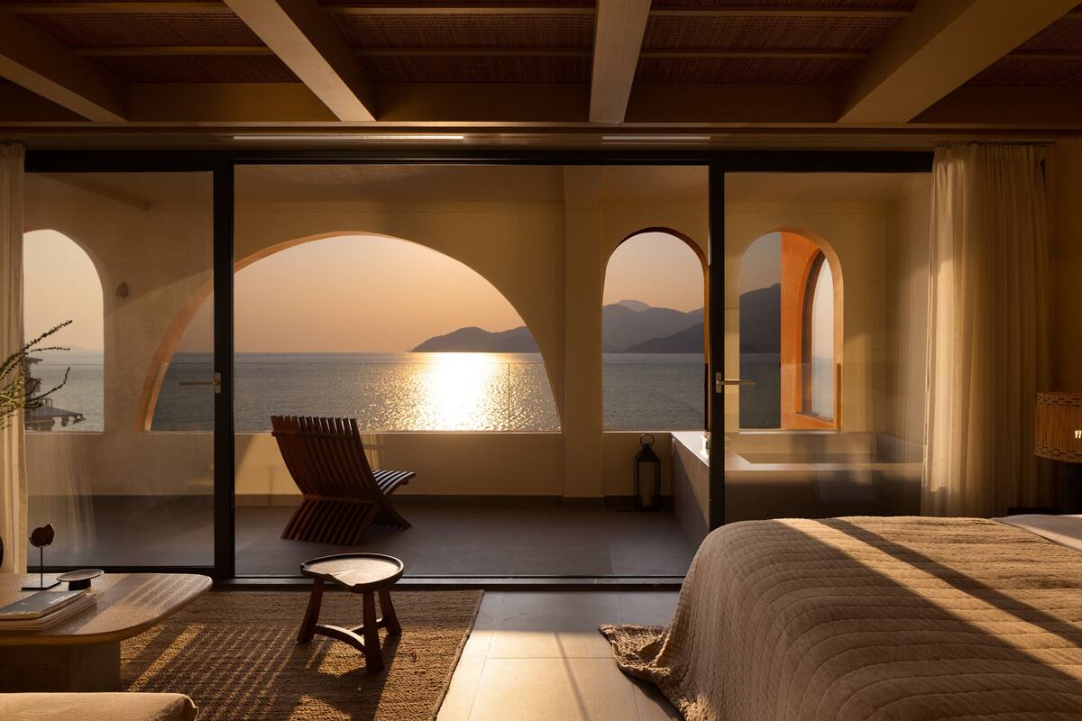 A bedroom with a large bed, a chair, and a view of the sea through arched windows.