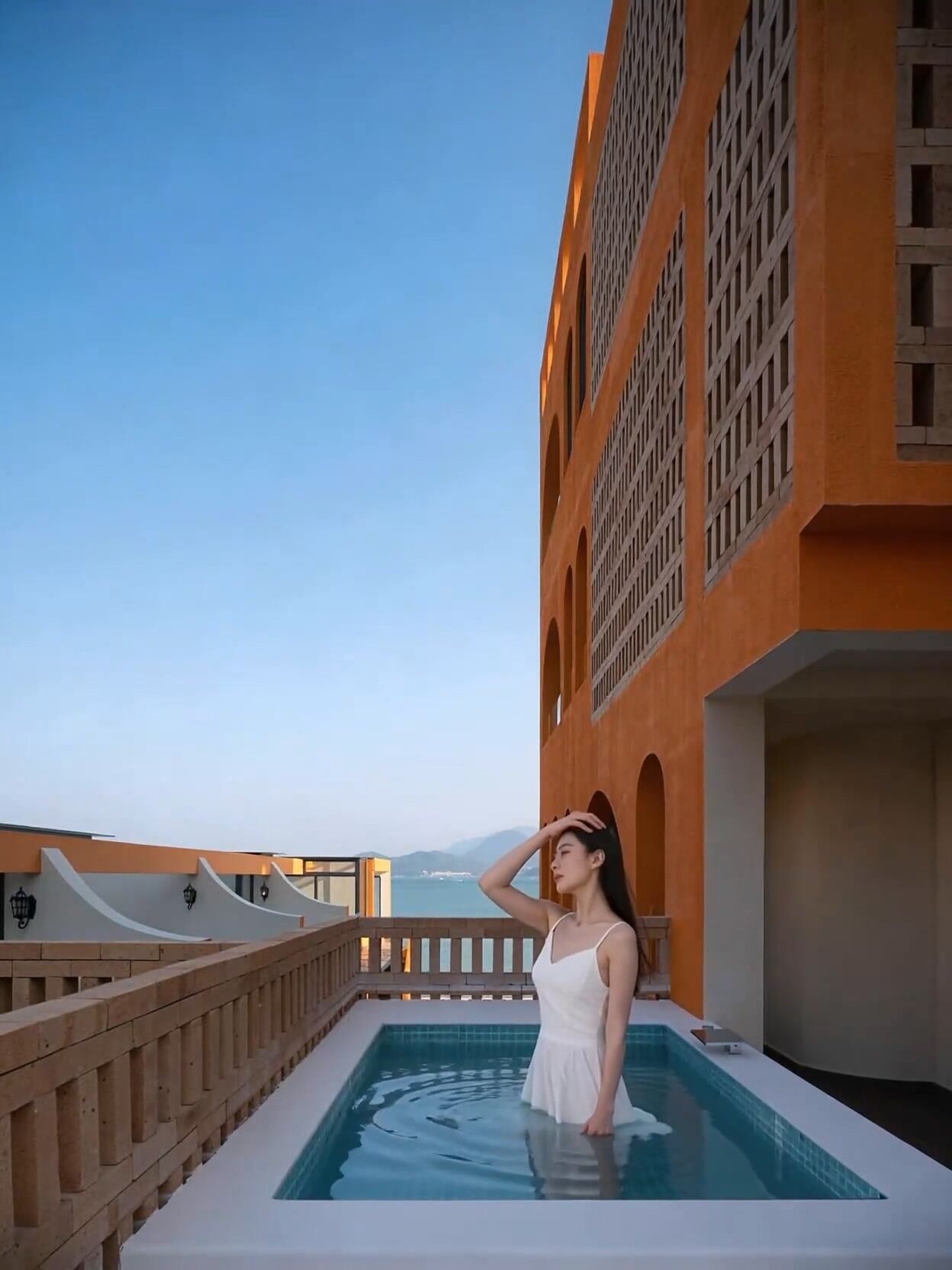 A woman in a white dress standing in a pool on a balcony with a view of the sea and mountains.
