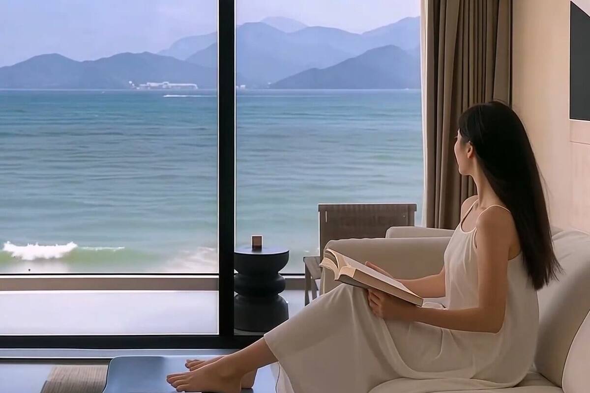 A room with a large window offering a view of the sea and mountains. A person is seated on a sofa reading a book.