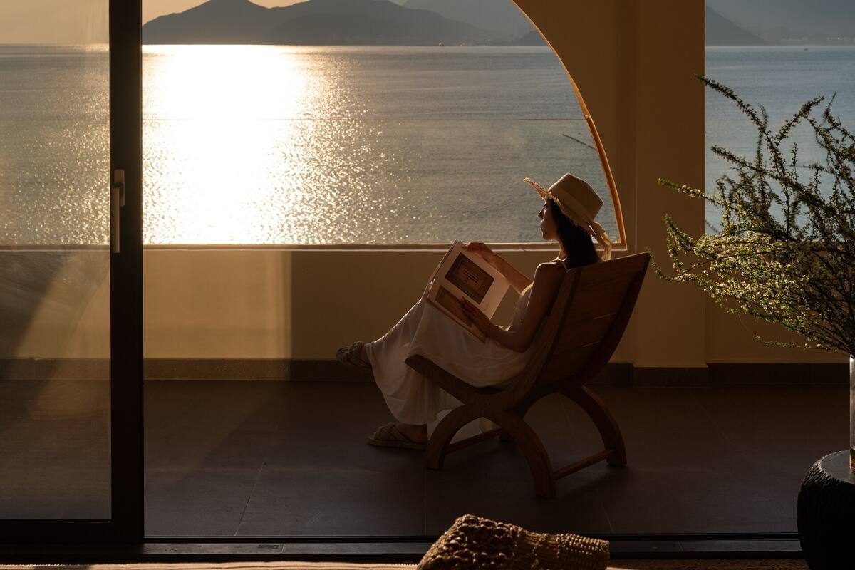 A room with a large arched window overlooking a body of water and mountains. There is a person reading a book, a chair, and a small table with a plant.