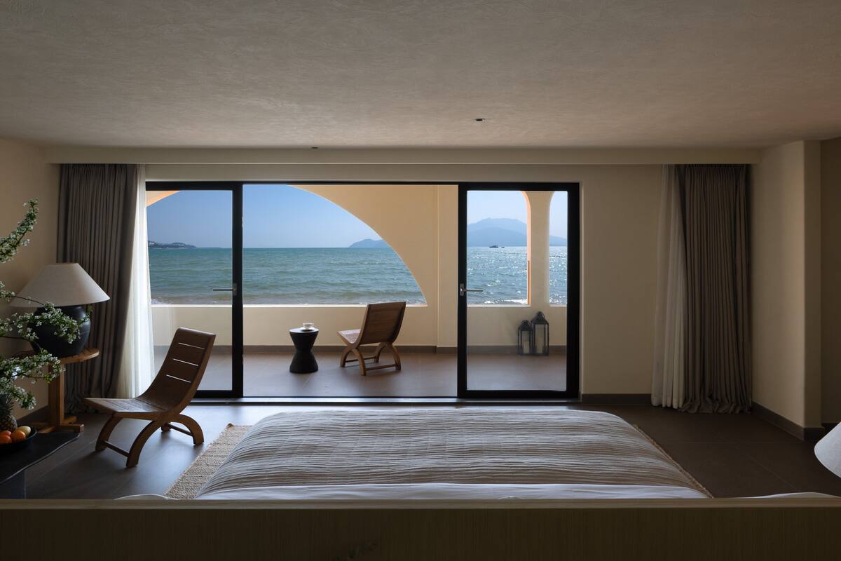 A hotel room with a large bed, a chair, a small table, and a view of the sea through a glass door.