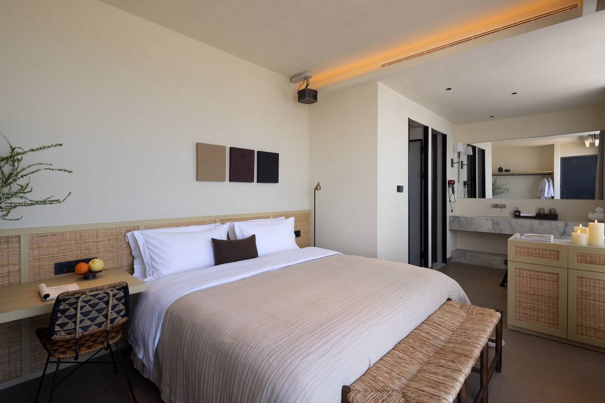 A hotel room with a large bed, a desk with a chair, a small table with fruits, a mirror, and a bathroom with a vanity and sink.