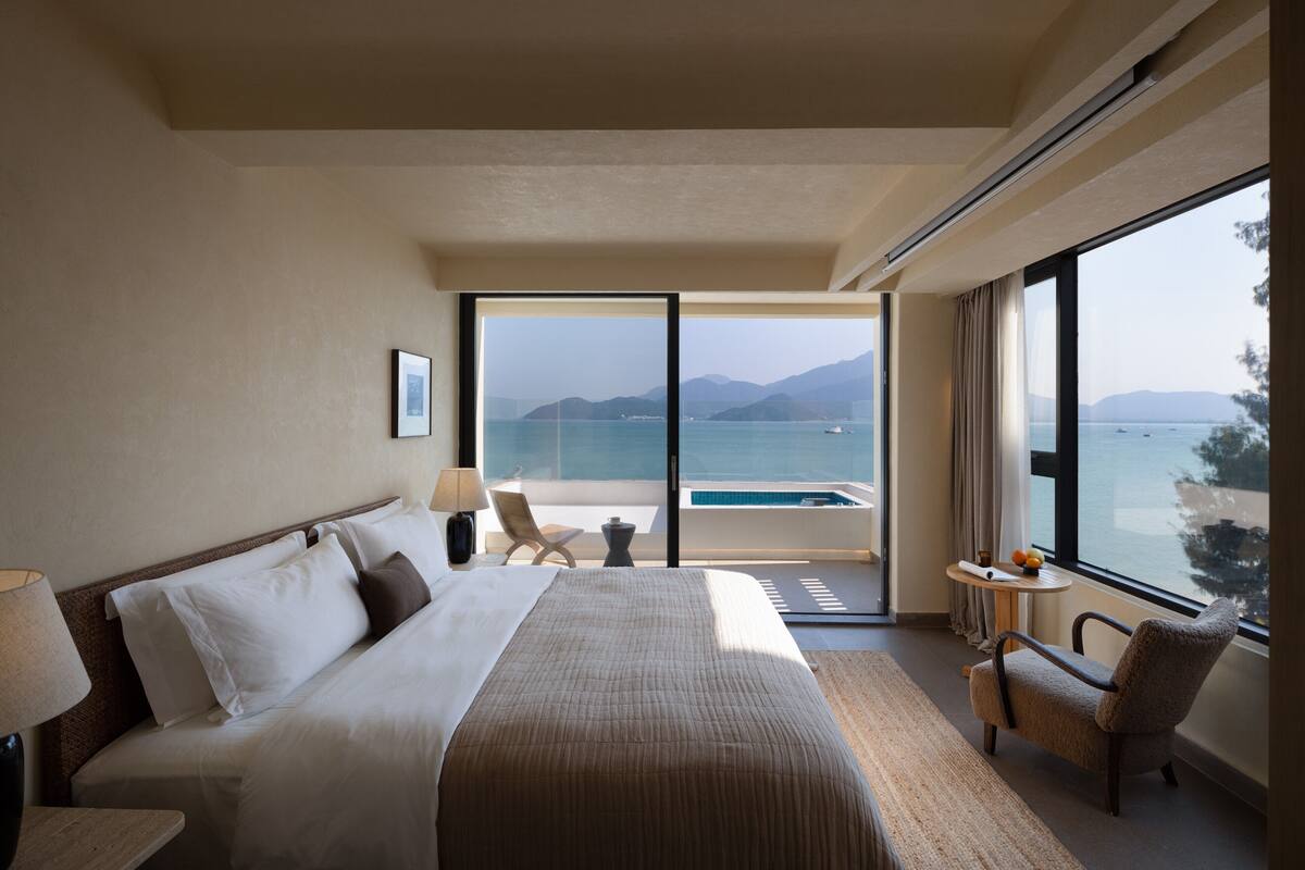 A hotel room with a large bed, a view of the sea, and a small table with a chair.