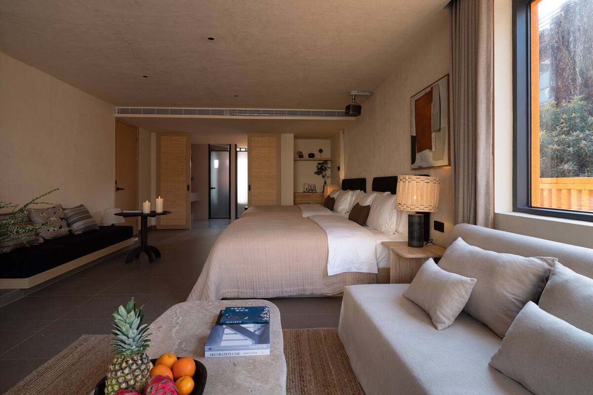 A hotel room with a large bed, a sofa, a coffee table with fruits and a book, and a window with a view of trees.