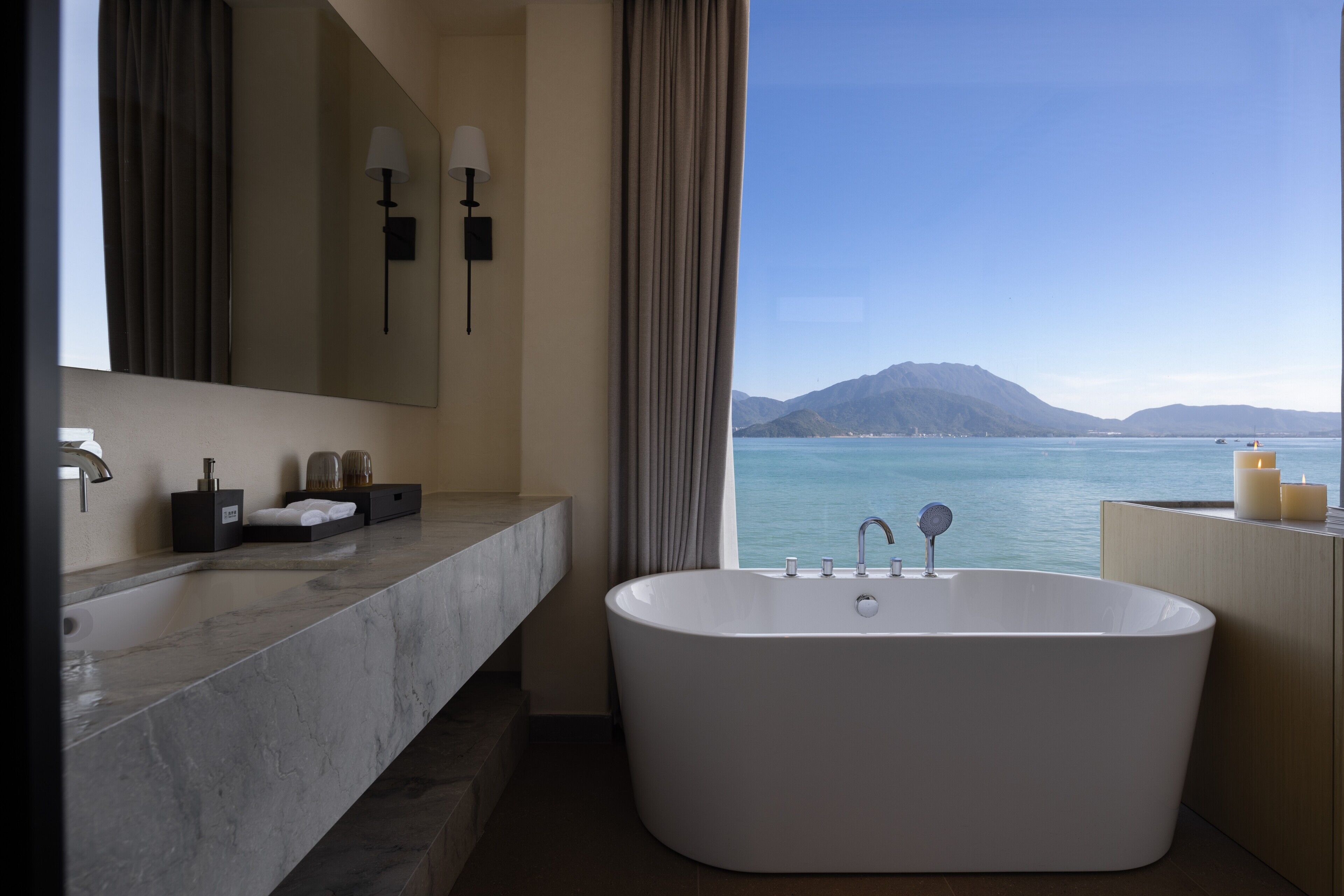 Une salle de bain moderne avec une grande baignoire, un meuble-lavabo en marbre, un grand miroir et une vue sur la mer.