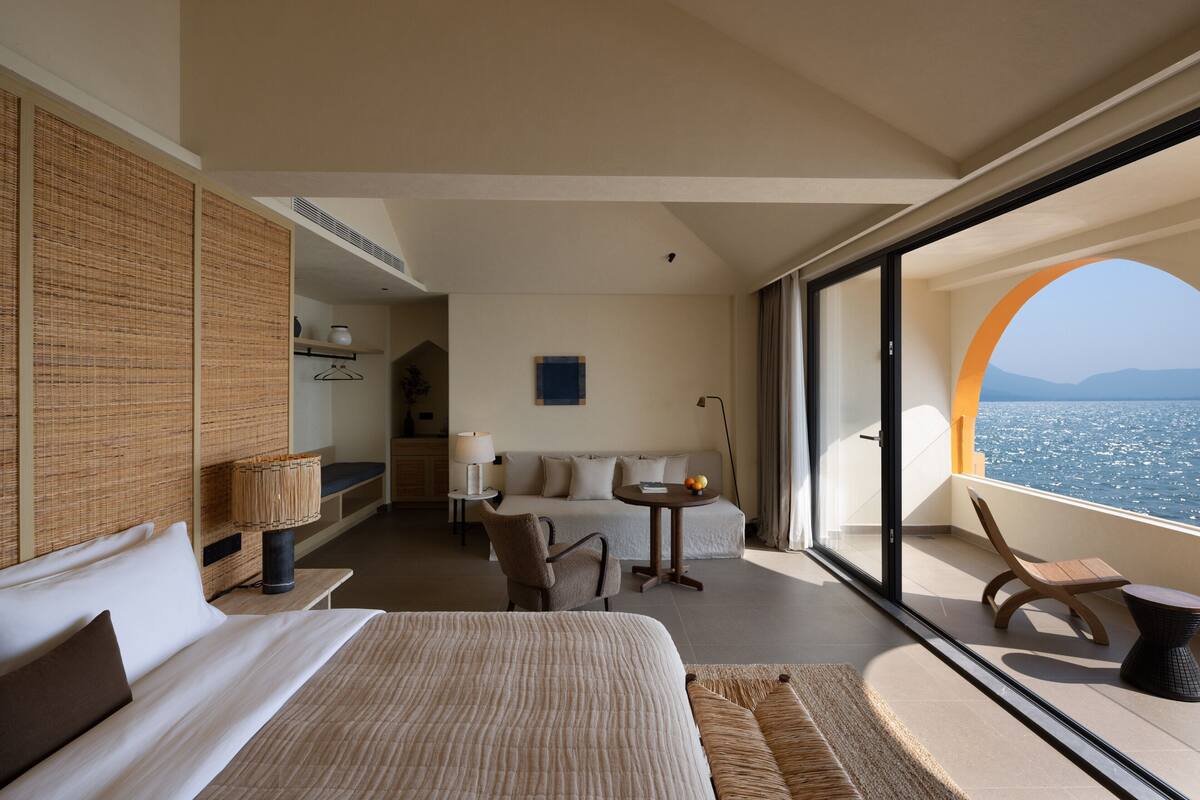 A hotel room with a large bed, a seating area with a table and chairs, a view of the sea through a glass door, and a balcony with a chair.