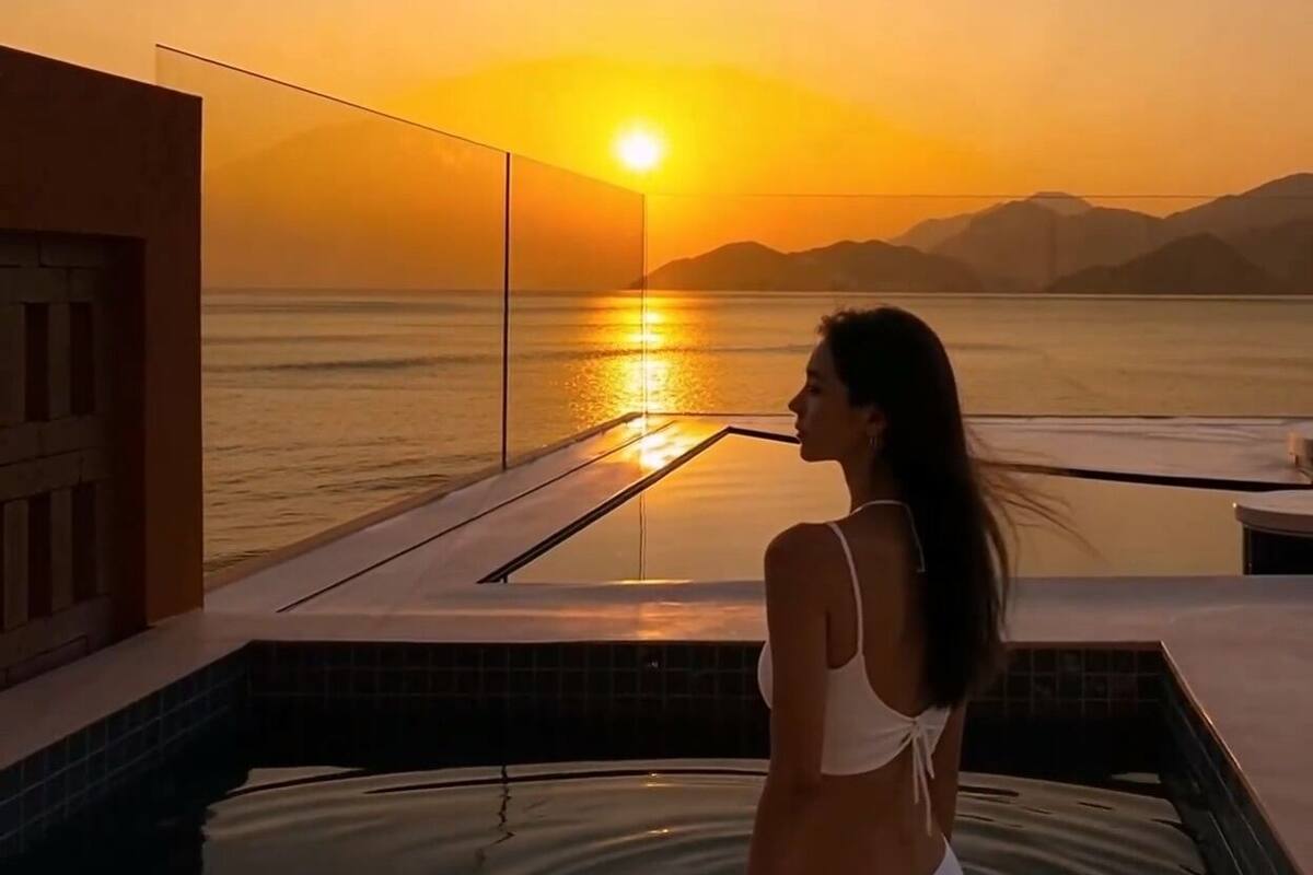 A person in a white swimsuit in a hot tub overlooking a sunset over water with mountains in the distance.