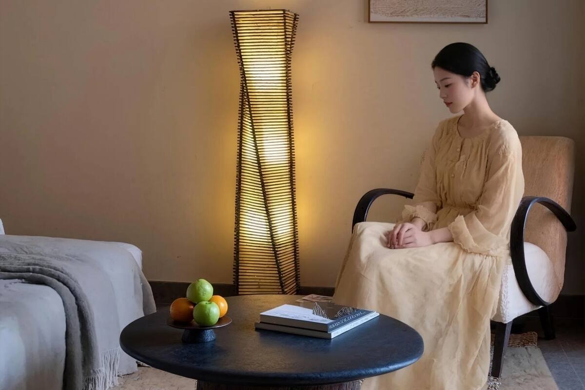 A room with a bed, a round coffee table with fruits and a book, a tall floor lamp, and a chair.