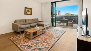 A modern living room with a sofa, coffee table, and a TV. There's a balcony with a view of a cityscape and a building.
