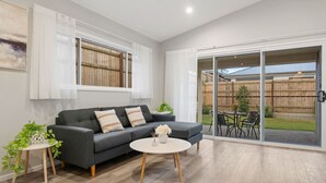 A modern living room with a grey sofa, two side tables, and a round coffee table. There is a sliding glass door leading to an outdoor patio area with a table and chairs. - Newly Built 4BR Family Oasis | Comfort & Space (Logan Reserve)