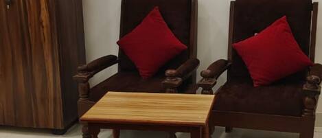 Two wooden armchairs with red cushions and a small wooden table in a room with a white wall and a wooden wardrobe.