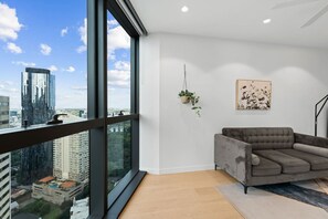 A modern living room with a large window, a grey sofa, a hanging plant, and a framed artwork on the wall.
