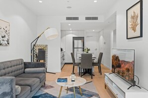 A modern living room with a sofa, coffee table, and TV. There is a dining area with chairs and a table. The room has white walls and a ceiling with recessed lighting.