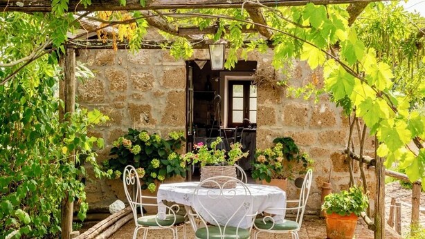 A stone building with a rustic outdoor seating area, including a table and chairs, surrounded by potted plants and vines. - La Cervaiola  (Porano)