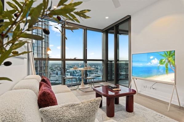 A modern living room with a city view, a dining table, and a television displaying a beach scene.
