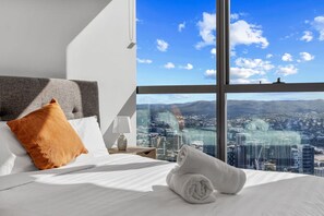 A hotel room with a large window offering a city view, a neatly made bed with white linens, a grey headboard, and a single orange pillow. - 59th Floor Luxury | Skyline & River Views (Brisbane City)