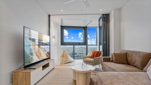 A modern living room with a large TV, comfortable seating, a coffee table, and a view of a cityscape through large windows.