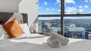 A hotel room with a large window offering a city view, a neatly made bed with white linens, a grey headboard, and a single orange pillow.