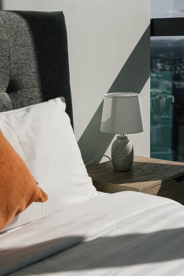 A bed with white bedding, a gray pillow, and a white lamp on a wooden nightstand.
