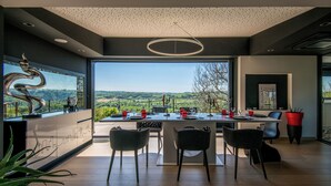 A modern dining area with a panoramic view, featuring a dining table set for four, surrounded by chairs, and a large window overlooking a scenic landscape. - New: Le Havre du Py, 230m² nestled in the heart of the fields (Compolibat)