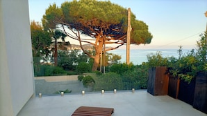 A balcony with a wooden bench, a glass railing, and a view of the sea and trees.