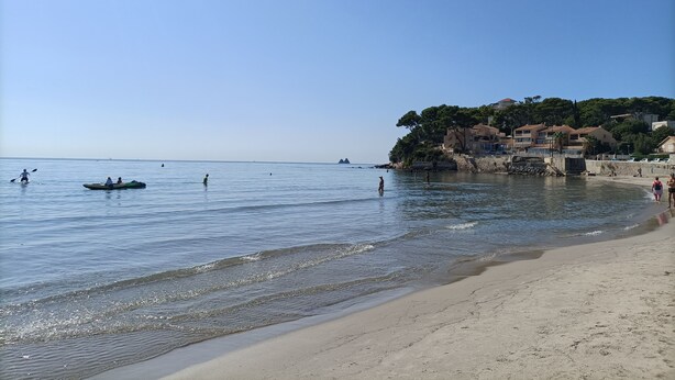 A beach with clear water, a sandy shore, and buildings in the background.