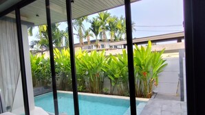 Una habitación moderna con un amplio ventanal corredero que da a una zona de piscina, rodeada de abundante vegetación y palmeras.
