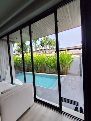A modern room with a large sliding glass door leading to a swimming pool area, surrounded by lush greenery and palm trees.