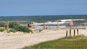 Een strand met een houten omheining en een bordje, een weg in de voorgrond en de oceaan op de achtergrond.