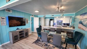 Een woonkamer met een televisie, een eettafel met glazen blad en teal-kleurige stoelen.