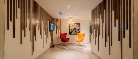 A modern hallway with a patterned floor, two chairs, a mounted TV, and a chandelier.