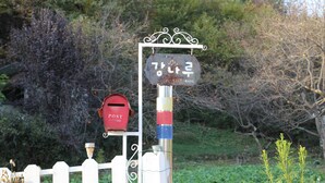 Un buzón sujeto a un poste de metal con un adorno en la parte superior, una valla de piquetes blancos y follaje al fondo.