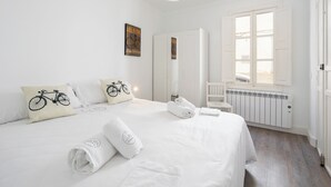 Une chambre d’hôtel avec un grand lit, du linge de lit blanc, un coussin à motifs de vélos, un miroir, un radiateur et une fenêtre avec des volets blancs.