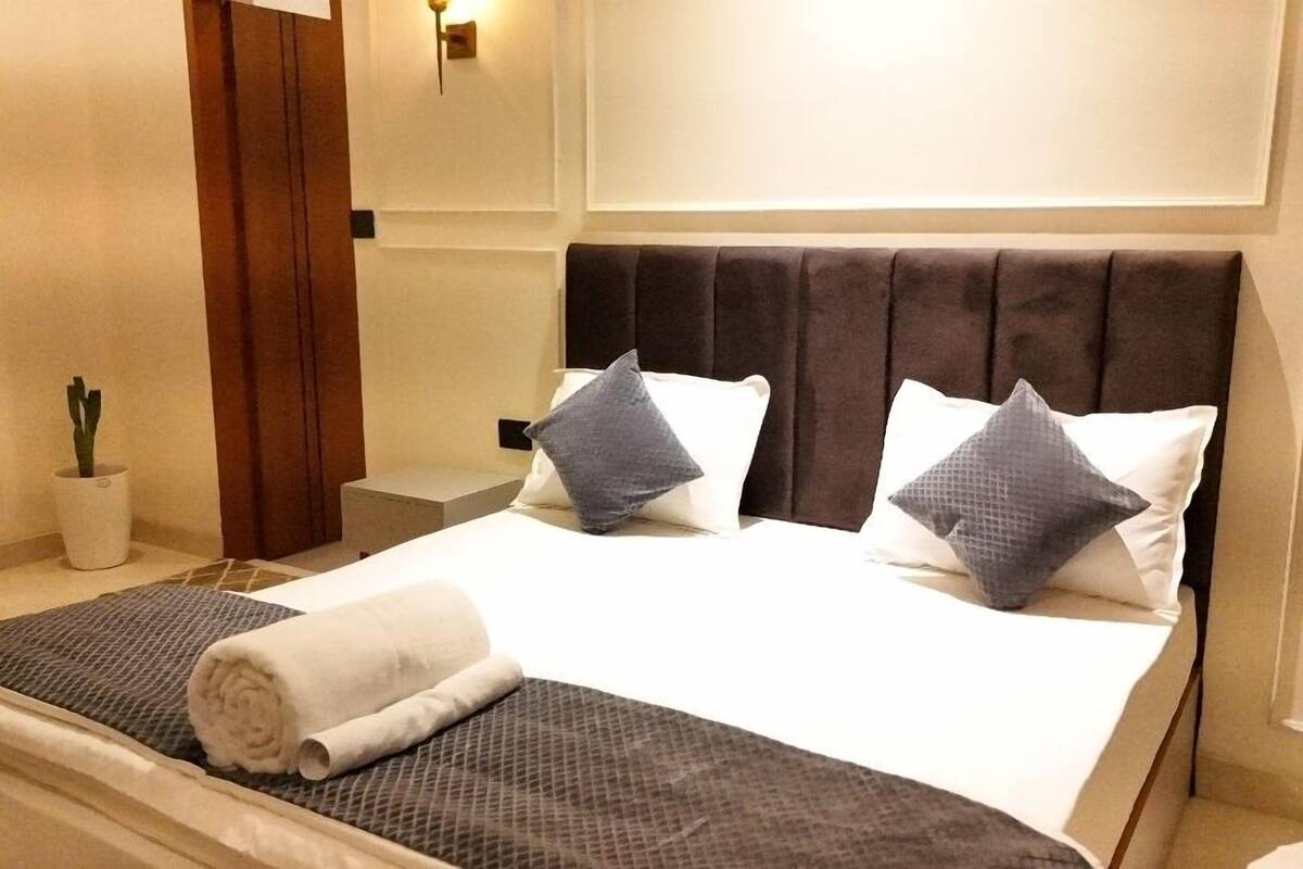 A hotel room with a large bed, two pillows, a bedside table, a nightstand, a potted plant, and a wall-mounted light fixture.
