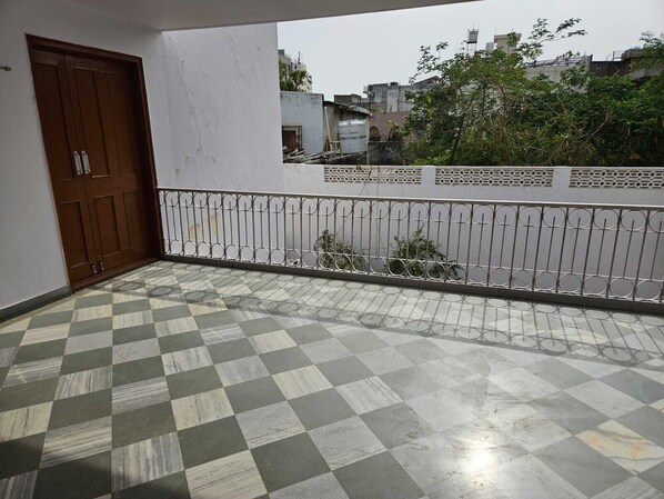 A balcony with a checkered floor, a metal railing, and a view of buildings and trees in the background. - Gullu Kuku Homestay (Jaipur)