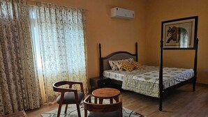 Une chambre à coucher comprenant un lit, une chaise, une petite table, une fenêtre avec des rideaux et un climatiseur fixé au mur.
