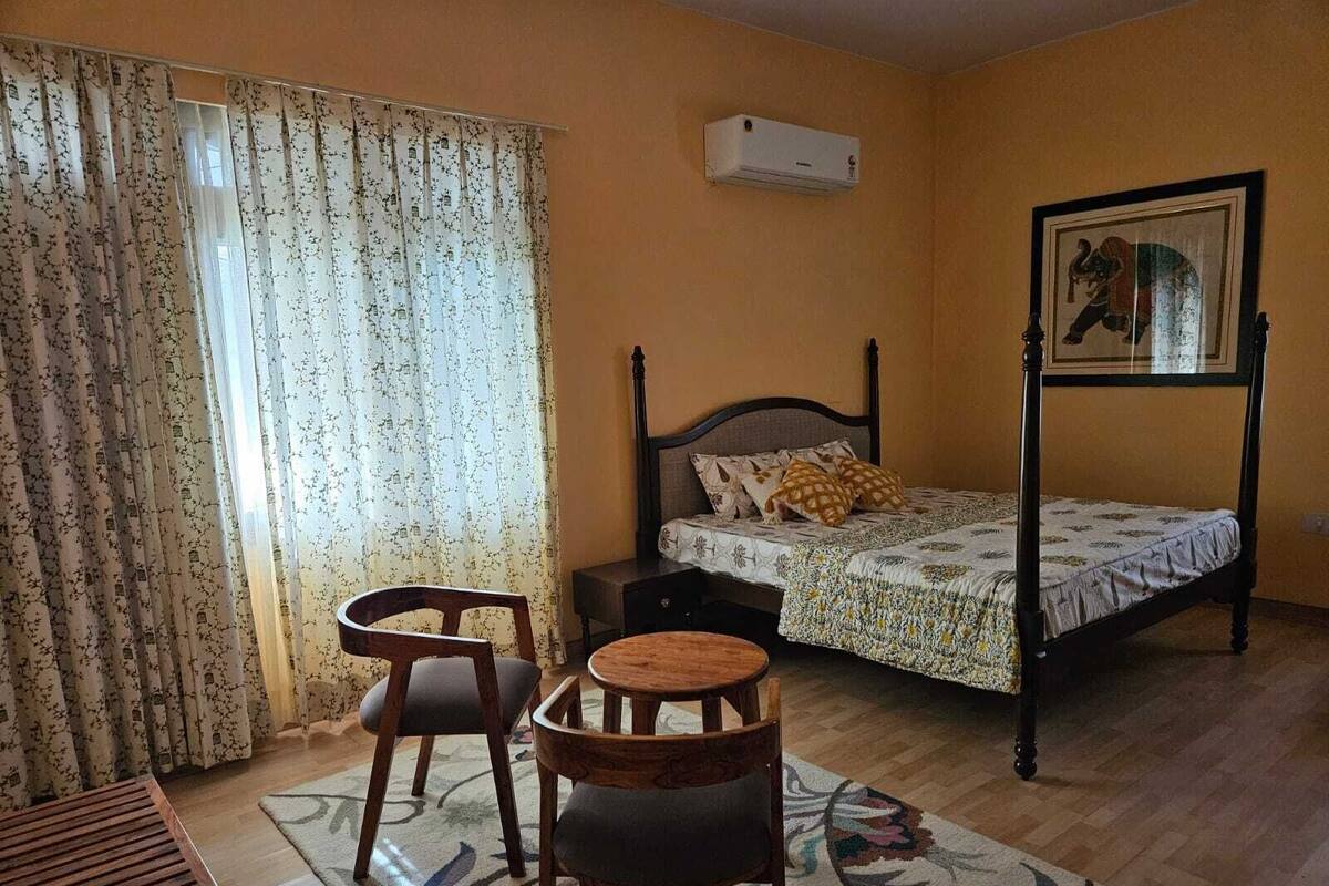 A bedroom with a bed, a chair, a small table, a window with curtains, and a wall-mounted air conditioner.