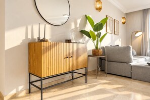 A modern living room with a wooden cabinet, a grey sofa, a round mirror, and a potted plant. - Sea & Sun Hills | Sea Views Pool (Estepona)