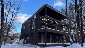 A modern two-story building with large windows and a flat roof, surrounded by snow-covered trees. - Forest Gardens (Hakuba)