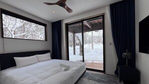 A modern bedroom with a large bed, a ceiling fan, a sliding glass door leading to a deck, and a view of a snowy landscape. - Forest Gardens (Hakuba)