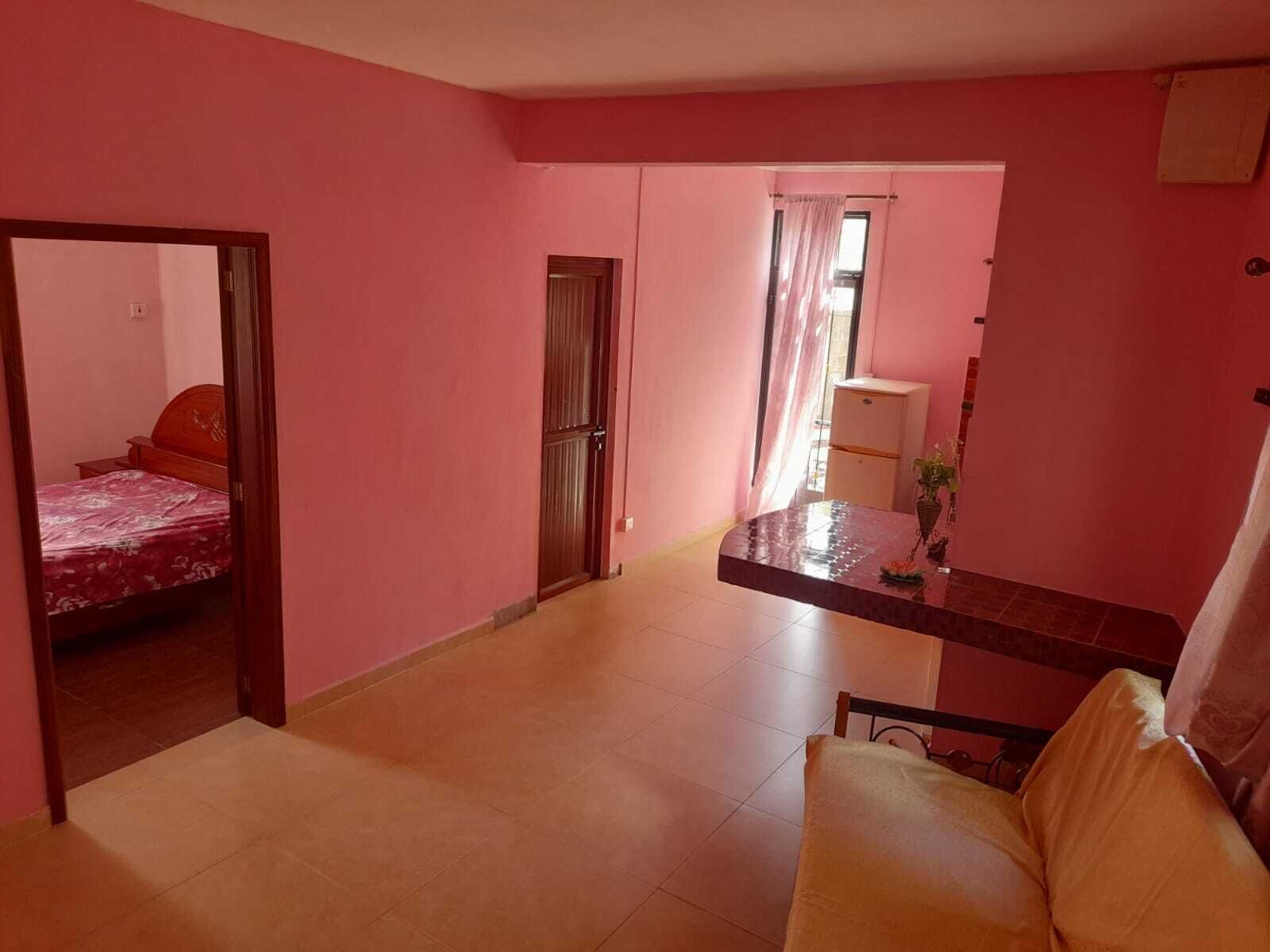 Una stanza dalle pareti rosa, con un letto e una coperta a motivi floreali, un tavolino con una pianta e una sedia.