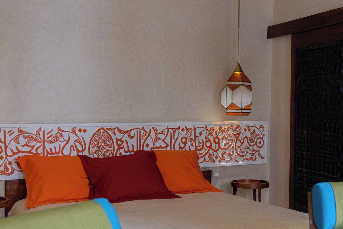 A hotel room with a bed, colorful pillows, a hanging light fixture, and a patterned floor.