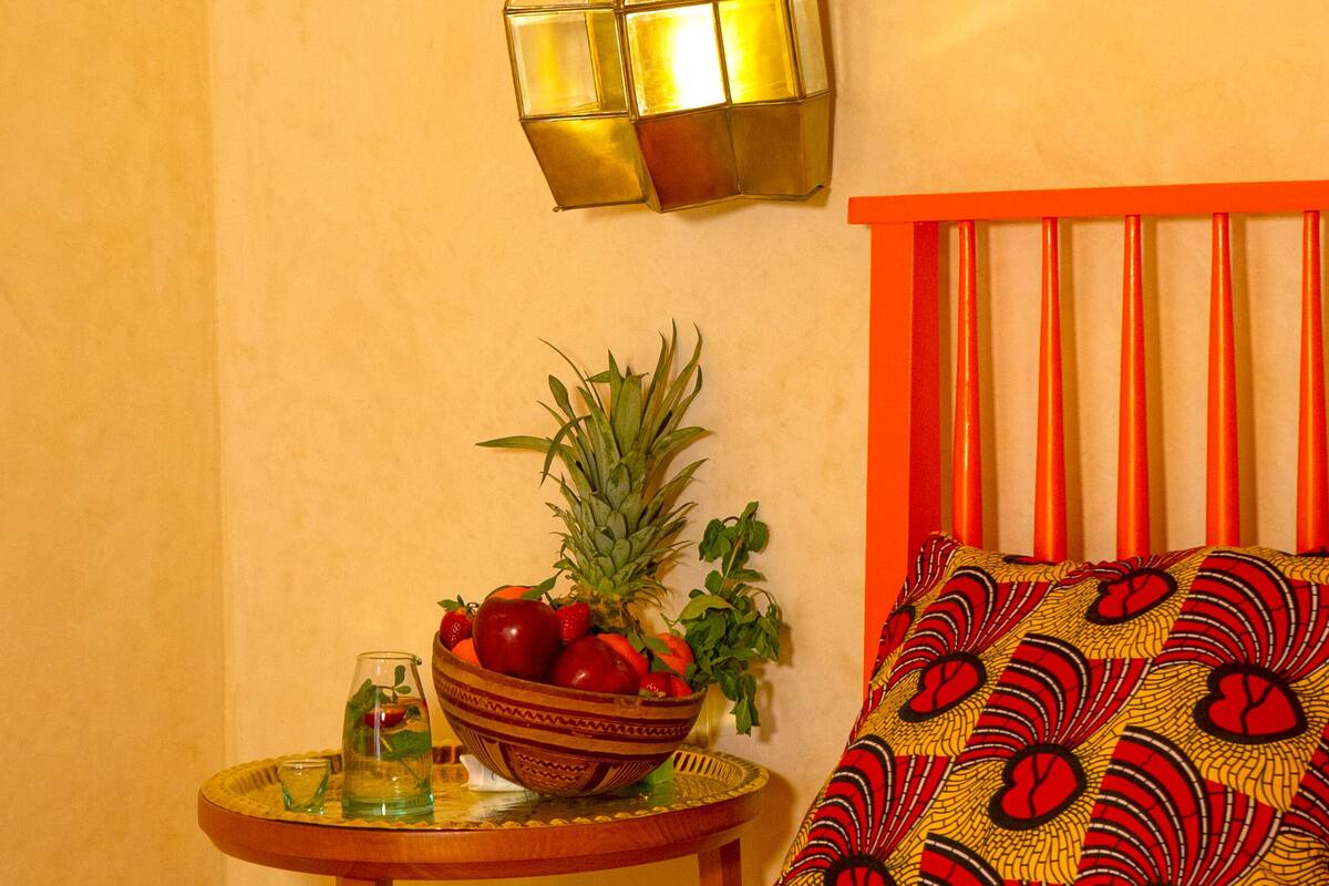 A bed with an orange headboard, a small round table with a bowl of fruit, a glass of water, and a green plant.